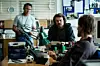 Tre mænd sidder og står omkring et rodet kontorskrivebord i seriøs dialog.