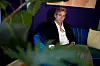 Mand i mørk jakke sidder på farverig sofa bag grønne planter i en stue