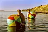 To voksne i havet med store, farverige strandbolde nær en kystklippe.