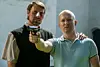 To mænd, hvor den ene sigter med en pistol og den anden står bagved med en hånd på skulderen.
