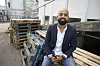 Mand i blazer og skjorte sidder smilende på træpaller i et industrielt gårdmiljø.