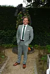 I jakkesæt foran en blomsterpyntet statue i en have med grøn hæk.
