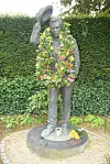 Bronzestatue med blomsterkrans foran tæt grøn hæk
