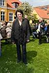 Pusle Helmuth står på en græsplæne ved et socialt arrangement foran gule huse.
