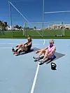 To kvinder laver siddende mavetræning med kettlebells på en udendørs sportsbane.