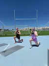 To personer laver lunges med kettlebell på en lys blå udendørs sportsbane i solskin.