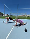 To kvinder træner planke med skulderklap på måtter på en udendørs bane med kettlebells omkring.