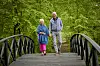 Hilda og Keld Heick går hånd i hånd over en træbro omgivet af grøn skov