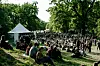 Stor menneskemængde sidder på græs i park med telte og træer ved udendørs arrangement.