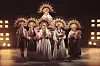 Fem performere i traditionelle dragter og blomsterkroner står på en oplyst scene med rekvisitter af korn.