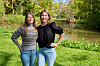 To kvinder står side om side på en græsplæne ved en sø i en grøn park.