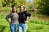 To kvinder står side om side på en græsplæne ved en sø i en grøn park.