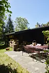 Terrasse med bord og stole ved sort træhus og store brændestabler i solskin