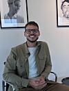 Jesper Alexander Blok Smilende med briller sidder foran væg med to indrammede portrætter.