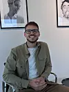 Jesper Alexander Blok Smilende med briller sidder foran væg med to indrammede portrætter.