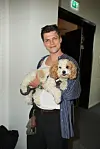 Holder en lille, lys krøllet hund på armen i en gang indendørs.