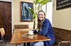 Sara Lygum sidder ved træbord på café med kaffekop og plante i baggrunden.