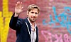 Ryan Gosling i jakkesæt hilser med løftet hånd foran mur med graffiti