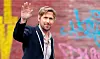 Ryan Gosling i jakkesæt hilser med løftet hånd foran mur med graffiti