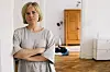 Person med korslagte arme står i en lys stue med trægulv og klædeskab bagved.