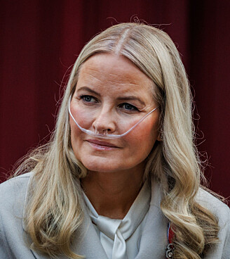 Kronprinsesse Mette-Marit sidder foran en mørkerød væg med næsekateter og lyse lokker.