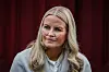 Kronprinsesse Mette-Marit sidder foran en mørkerød væg med næsekateter og lyse lokker.