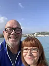 Smilende par med briller foran havn og kystby i klart sollys.