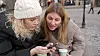 To kvinder i vintertøj deler en smartphone ved et udendørs cafébord