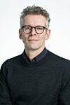 Portræt af mand med briller i mørk trøje foran lys baggrund.