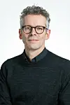 Portræt af mand med briller i mørk trøje foran lys baggrund.