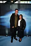 Et smilende par poserer foran en blå oplyst fotovæg ved en filmpremiere.