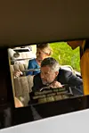 Kong Frederik kigger ud af vinduet i den røde Bedford-bus, der kører forbi 'Badehotellet'.