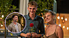 To unge personer smiler med en rød rose og champagneglas ved et udendørs aftenarrangement.