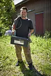 Smilende Simon i arbejdstøj bærer metalboks og kabel foran rød træbygning i solskin