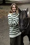 Smilende kvinde i striktrøje står tæt ved en mørk hest på en skovsti.