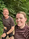 To smilende løbere i brune trøjer løber på skovsti omgivet af grønne træer.