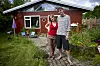 To personer står på en stensti foran et rødt træhus omgivet af grøn sommerhave.