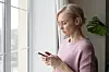 Ældre kvinde i rosa trøje står ved et vindue og ser bekymret på sin smartphone.
