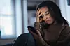 Kvinde sidder alene og græder, mens hun læser en besked på sin smartphone.