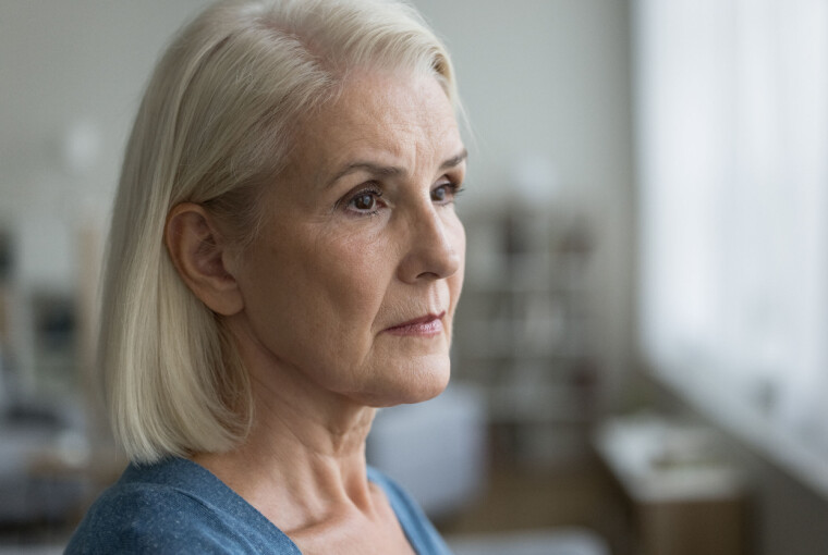 Tankefuld ældre kvinde med gråt hår kigger ud ad et lyst vindue indendørs.