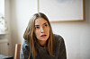 Ung kvinde i grå sweater sidder indendørs og ser tænksom ud.