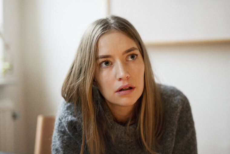 Ung kvinde i grå sweater sidder indendørs og ser tænksom ud.