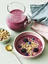 Skål med pink smoothiebowl toppet med granola, blåbær og brombær ved siden af kande smoothie.