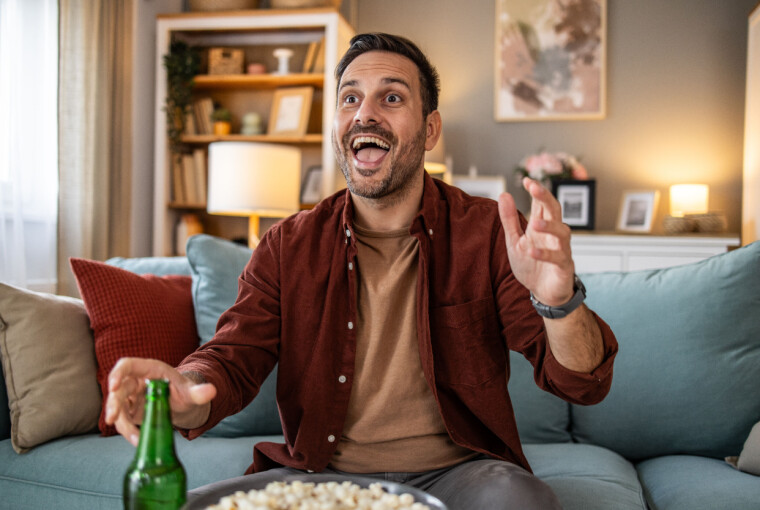 Mand på sofa reagerer begejstret på fodboldkamp med popcorn og øl foran sig.