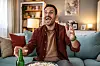 Mand på sofa reagerer begejstret på fodboldkamp med popcorn og øl foran sig.