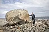 Mand i jakkesæt står ved stor kampesten på stenet kyst med hav i baggrunden