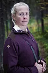 Kvinde i mørkelilla trøje står i skov og ser direkte mod kameraet