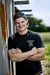Jonathan Nielsen står smilende med korslagte arme op ad en rusten ladevæg på landet.