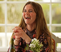Smilende kvinde ved bord med flasker og blomster i lyst rum