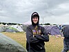 Person i hættetrøje står smilende på mudret festivalcampingplads omgivet af telte.